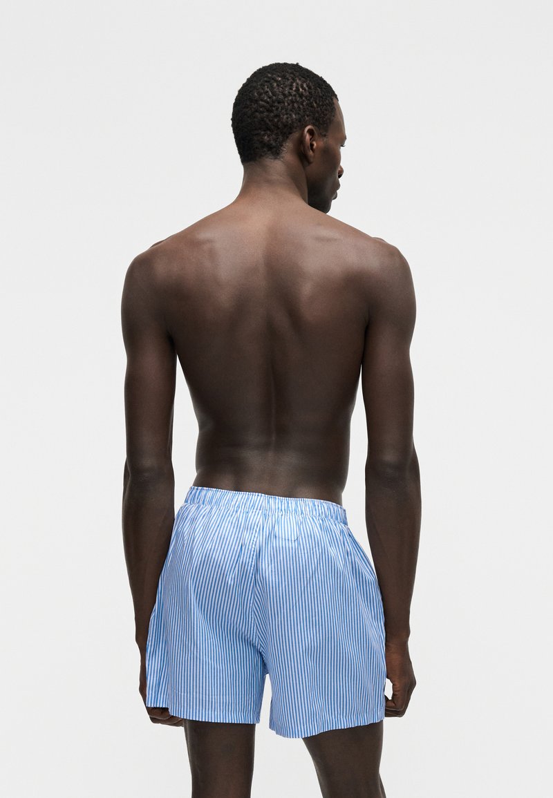 Man met een donkere huid die van achteren te zien is, gekleed in lichtblauwe en witte verticaal gestreepte korte broek tegen een effen lichte achtergrond.