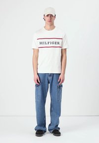 White cotton t-shirt with red and navy stripe text "HILFIGER," paired with blue denim cargo pants and black slip-on shoes. Light cap.