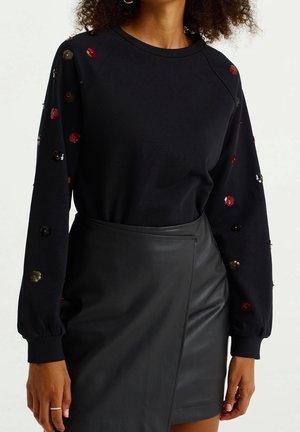 Mujer con una camiseta negra de manga larga con flores multicolores de lentejuelas y una falda negra de cuero tipo pareo.