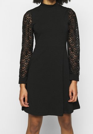Femme portant une robe noire à col montant et longues manches en dentelle transparente à motifs, debout devant un fond blanc.