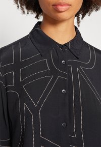 Maglietta nera con un design a bottoni, caratterizzata da cuciture bianche in contrasto con motivi geometrici e un colletto classico. Texture liscia.