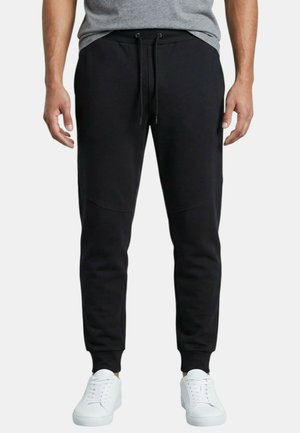 Homme portant un pantalon de jogging noir avec taille à cordon et des baskets blanches, debout devant un fond uni.