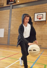 Schwarze Zip-Hoodie über einem weißen T-Shirt, schwarze Hose und weiße Sneaker. Ein Basketball wird auf einem Holzboden mit bunten Linien gedribbelt.