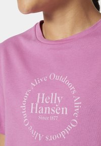 Růžové bavlněné tričko s kulatým bílým potiskem, na kterém je nápis "Helly Hansen Since 1877" a "Alive Outdoors" v kruhovém textovém vzoru.