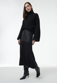 Suéter negro de gran tamaño con mangas anchas, combinado con una falda midi negra de canalé. La modelo lleva botas de cuero negro de punta con altura hasta la rodilla.