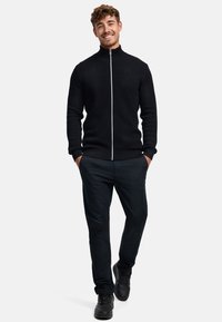 Musta vetoketjullinen neulepusero, jossa on teksturoitu kuvio, korkea kaulus ja kontrastinen valkoinen vetoketju. Yhdistetty tummiin housuihin ja mustiin tennareihin.