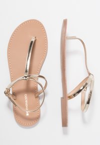 Platta sandaler med ett metalliskt guldremdesign, en beige fotbädd och en texturerad gummisula. Enkel, minimalistisch stil med ankelremmar.