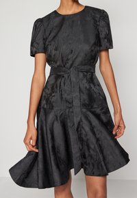 Robe noire avec des manches bouffantes courtes, un corsage ajusté, une jupe évasée et une ceinture en tissu assorti à la taille. Fini texturé.