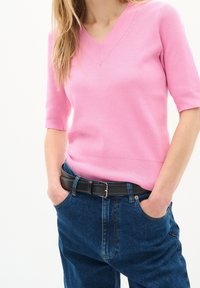 Růžový pletený svetr s V-výstřihem a krátkými rukávy, kombinovaný s tmavě modrými džínami a černým opaskem.