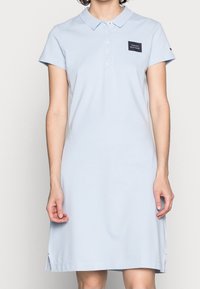 Světle modré polo šaty vyrobené z bavlněné směsi. Mají límeček, krátké rukávy a malou černou nášivku na hrudi. Po stranách mají rozparky.