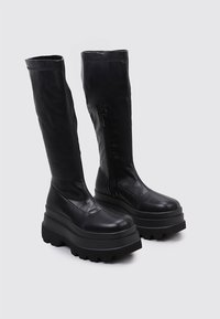 Botas de plataforma de cuero negro hasta la rodilla con suela gruesa, punta redondeada y cremallera lateral externa para facilitar su uso. Textura suave.