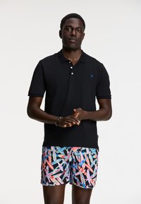 Čierne polo tričko s tromi bielymi gomb�íkmi a modrým emblémom, spárované s mnohofarebnými šortkami s abstraktnými vzormi.