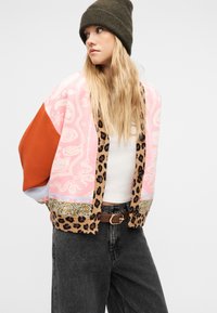 Γυναίκα με πράσινο ελαιοπράσινο σκουφί, φαρδιά ροζ ζακέτα με λεοπάρ σχέδιο, λευκό τοπ, καφέ ζώνη και μαύρο τζιν, κοιτώντας προς τα πλάγια.