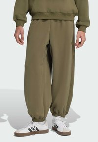 Pantalones de chándal verde oliva de tejido suave con puños elásticos, con corte holgado y bolsillos laterales. Combinados con zapatillas blancas con rayas negras.