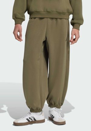 Olivegrüne Sweatpants aus weichem Stoff mit elastischen Bündchen, lockerer Passform und seitlichen Taschen. Kombiniert mit weißen Sneakers mit schwarzen Streifen.