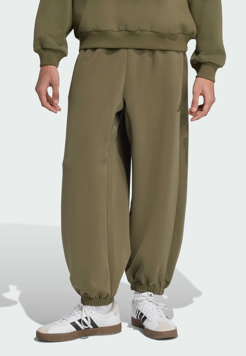 Pantalones de chándal verde oliva de tejido suave con puños elásticos, con corte holgado y bolsillos laterales. Combinados con zapatillas blancas con rayas negras.