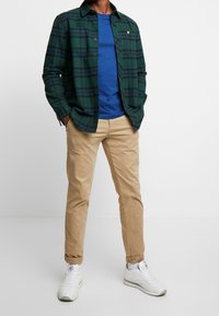 Camisa de cuadros verde y marino con botones sobre una camiseta azul, combinada con pantalones chinos beige y zapatillas blancas. Detalle del logo dorado en la camisa.