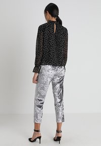 Schwarze, durchsichtige Bluse mit kleinen weißen Blumenmustern und langen Ärmeln, kombiniert mit glänzenden, silbernen Samt-Hosen in Knöchelhöhe.