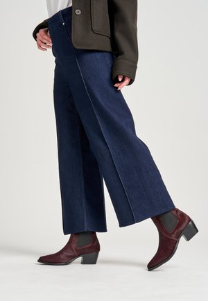 Pantaloni in denim a gamba larga di colore blu scuro con una silhouette dritta, abbinati a stivaletti borgogna con pannelli testurizzati e tacco basso.