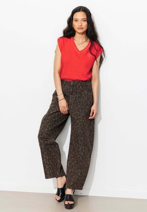 Femme aux longs cheveux foncés portant un haut rouge sans manches avec un petit détail en forme de cœur, un pantalon large marron à imprimé léopard et des sandales compensées noires.