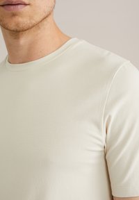 Tricou bej cu mânecă scurtă, realizat dintr-un material neted și ușor; are un guler clasic rotund și o siluetă ajustată.