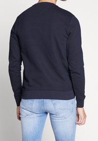 Námořnická modrá mikina s dlouhými rukávy, kulatým výstřihem a žebrovanými manžetami. Nošena s světle modrými slim fit džíny, ukazující neformální styl.