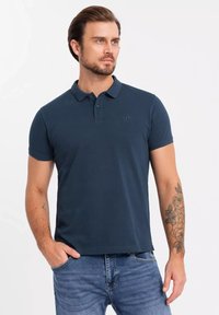 Mörkblå pikétröja i bomull med krage, tre-knapps knäppning och en diskret broderad logotyp på bröstet. Kombinerad med jeans.