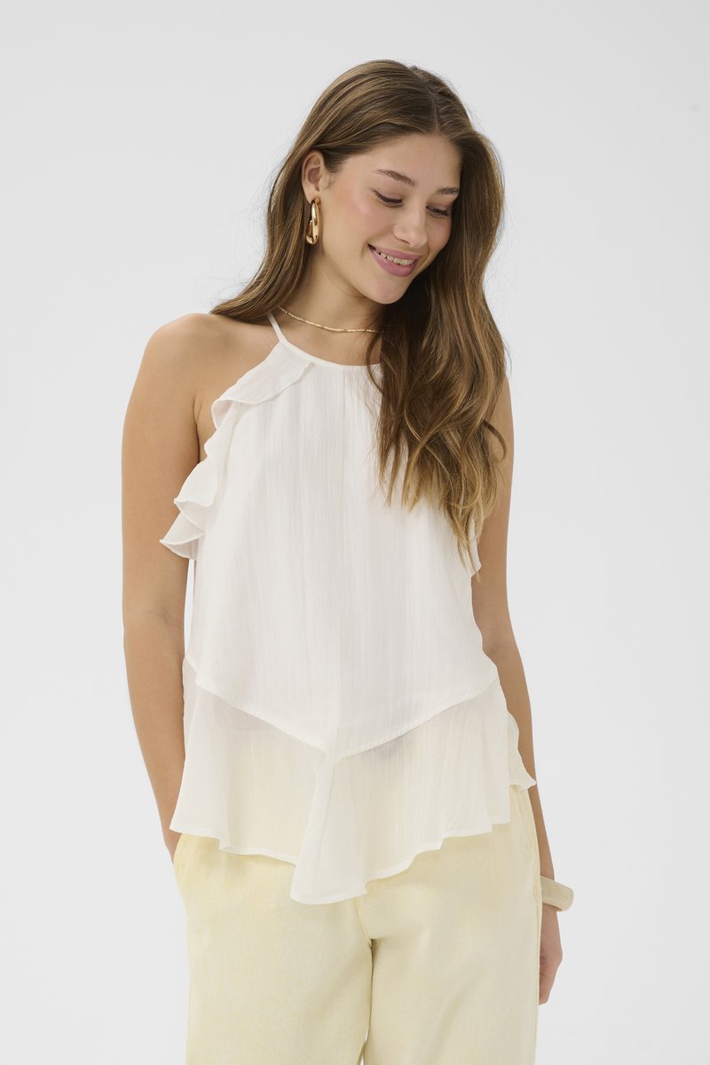 Femme souriante aux longs cheveux bruns portant des boucles d'oreilles créoles dorées, un haut blanc sans manches à volants et un pantalon clair, les mains dans les poches.