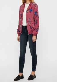 Blouson aviateur rose avec broderie florale en bleu et blanc, associé à un t-shirt blanc et un jean skinny foncé. Des mocassins noirs complètent le look.