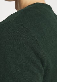 Ciemnozielony sweter z dzianiny o gładkiej fakturze, z okrągłym dekoltem i widocznymi szwami na ramionach.