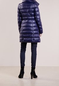 Marineblå pufferjakke med en skinnende finish, der har vandrette syninger, en høj krave og en tætsiddende silhuet, kombineret med mørke jeans og sorte støvler med hæl.