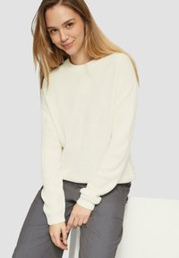 Witte gebreide trui met lange mouwen en geribbelde textuur, gecombineerd met grijze broek. Het model heeft steil haar en zit tegen een neutrale achtergrond.
