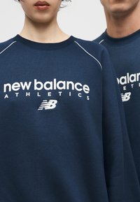 Δύο άτομα φορούν αθλητικές μπλούζες New Balance σε σκούρο μπλε χρώμα, με εμφανές το λογότυπο μπροστά και λεπτομέρειες στη γραμμή λαιμού.