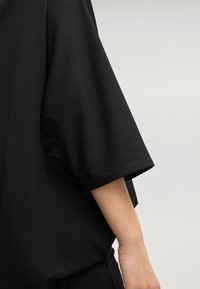 T-shirt noir avec une coupe ample et des manches larges et retroussées. Fabriqué en tissu léger et lisse avec une texture douce. Design minimaliste.