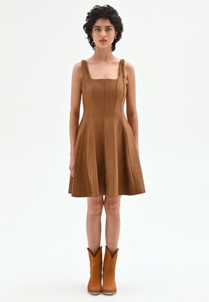 Femme aux cheveux courts et bouclés, portant une robe marron sans manches et des bottines marron assorties, debout droite sur un fond blanc uni.