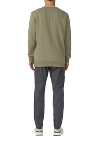 Uomo che indossa un pullover a maniche lunghe verde oliva, pantaloni cargo grigi con tasche laterali e sneaker bianche, in piedi di spalle su sfondo bianco.