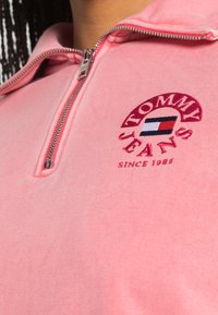 Felpa rosa con zip e texture a coste. Presenta il logo ricamato "Tommy Jeans" e un piccolo dettaglio a bandiera sul petto.
