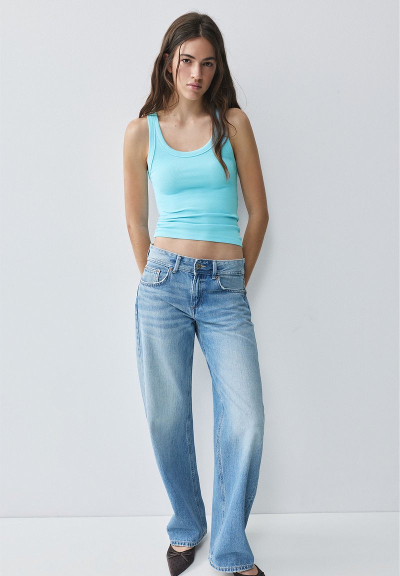 Jeune femme debout, mains dans le dos, portant un débardeur turquoise, un jean bleu clair et des ballerines foncées sur un fond uni.
