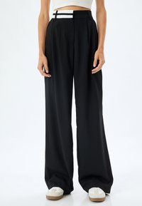 Pantalons larges noirs avec des plis et une ceinture distinctive à rayures blanches. Le matériau semble lisse, avec une coupe ample et un design taille haute.