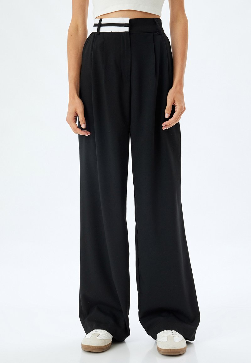 Pantalons larges noirs avec des plis et une ceinture distinctive à rayures blanches. Le matériau semble lisse, avec une coupe ample et un design taille haute.
