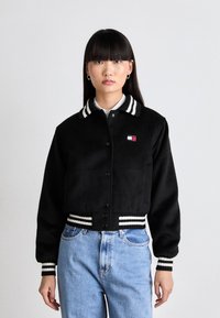 Sort cropped jakke med en blød tekstur, der har en stribet krave og manchetter. Viser et lille, farvet logo på brystet.