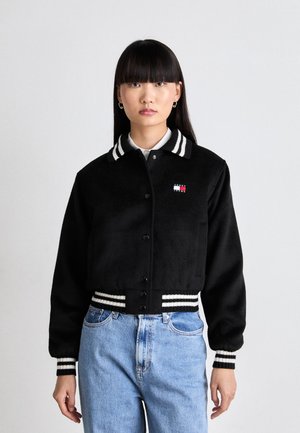 Schwarze, kurz geschnittene Jacke mit flauschiger Textur, ausgestattet mit einem gestreiften Kragen und Bündchen. Zeigt ein kleines, farbiges Logo auf der Brust.