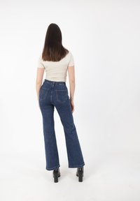 Donna con lunghi capelli castani che sta in piedi di spalle, indossando una maglietta beige a maniche corte, jeans blu a vita alta e stivali neri con tacco.