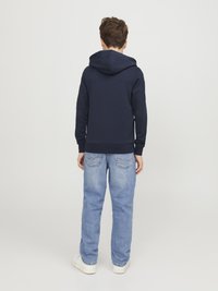 Felpa blu marina con tasca frontale e cappuccio regolabile, abbinata a jeans in denim blu chiaro e sneakers bianche. Design semplice, vestibilità casual.