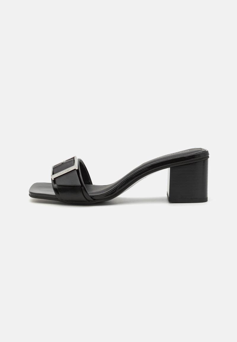 Mule noire à bout ouvert avec talon bloc et large bride avec une boucle rectangulaire argentée sur fond blanc.