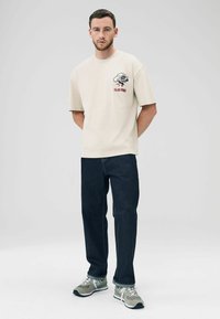 T-shirt en coton beige clair à manches courtes avec un imprimé graphique sur la poitrine, associé à un jean bleu foncé et des baskets grises.