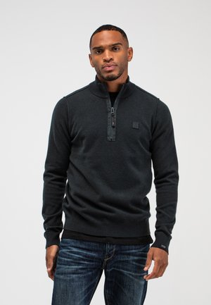 Man met kort haar en een verzorgde baard, gekleed in een donker half-ritssluitingtrui en blauwe spijkerbroek, staand tegen een effen witte achtergrond.