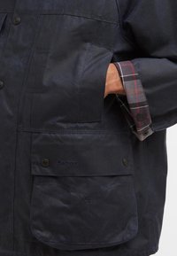 Casaco de algodão encerado azul-marinho com forro de algodão xadrez, apresentando dois grandes bolsos frontais e fechos de botão. O logotipo é visível no bolso.