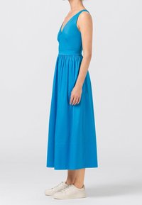 Blå ärmlös klänning med V-ringning, figurnära liv och en draperad, ankel-lång kjol. Bärs med ljusa sneakers.