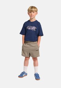 Marinfärgad grafisk t-shirt med lila tryck, beiga shorts, blå sneakers med vita detaljer och vita strumpor. Barnet står med händerna i shortsens fickor.